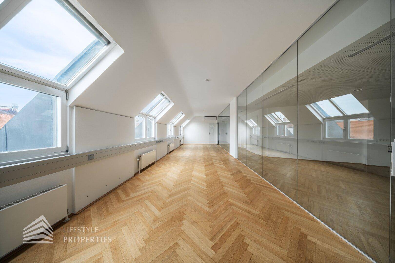 Langer Bürogang mit Glaswänden und Fischgrätparkett, der zu weiteren hellen Arbeitsbereichen im Dachgeschoss führt.