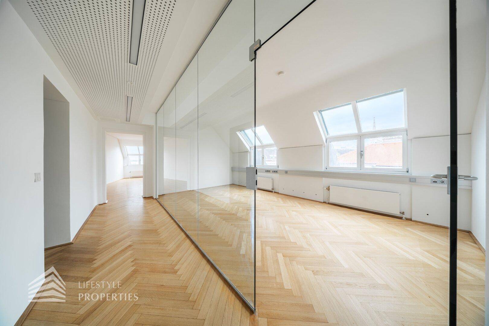 Moderner Büroraum mit Glasabtrennungen, hellem Fischgrätparkett und viel Tageslicht durch die Dachfenster.