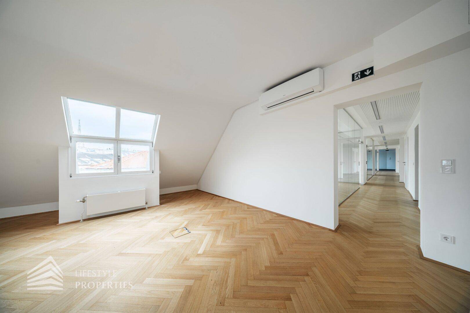 Heller Büroraum mit schräger Wand, Klimaanlage und Blick auf den Flur durch eine Glaswand.