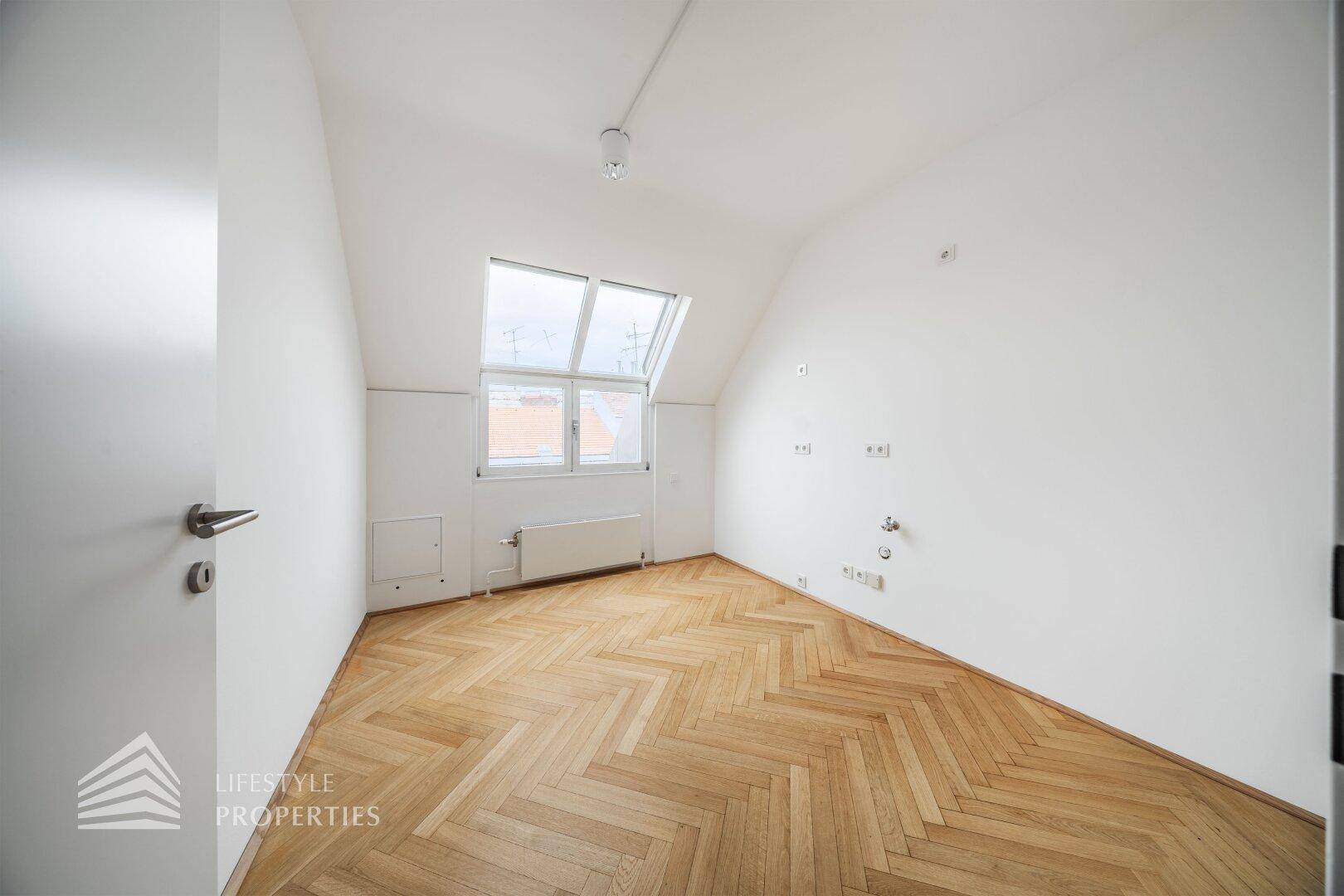 Leerer Büroraum mit Fischgrätparkett, Dachfenster und Heizkörper unter dem Fenster.