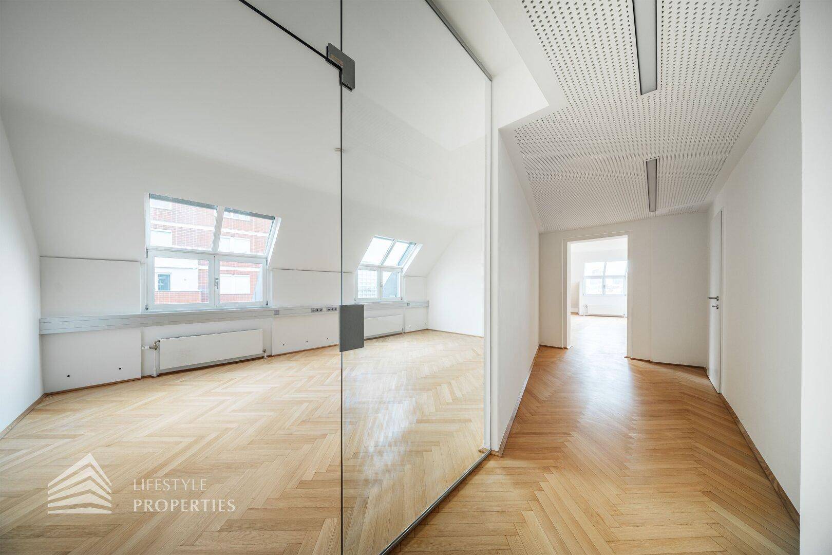 Bürobereich mit Glaswand, Fischgrätparkett und schrägen Fenstern, bietet eine helle und moderne Arbeitsumgebung.