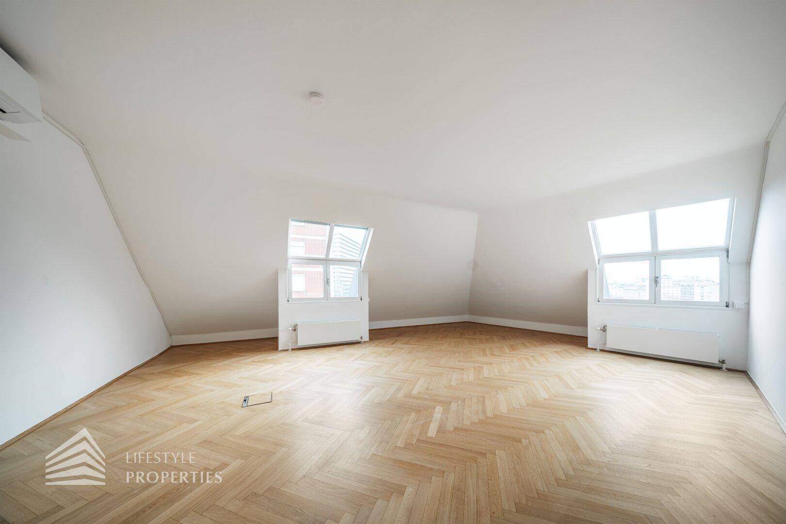 Heller Büroraum mit zwei schrägen Fenstern, Fischgrätparkett und weißer Decke, bietet eine offene und luftige Atmosphäre.