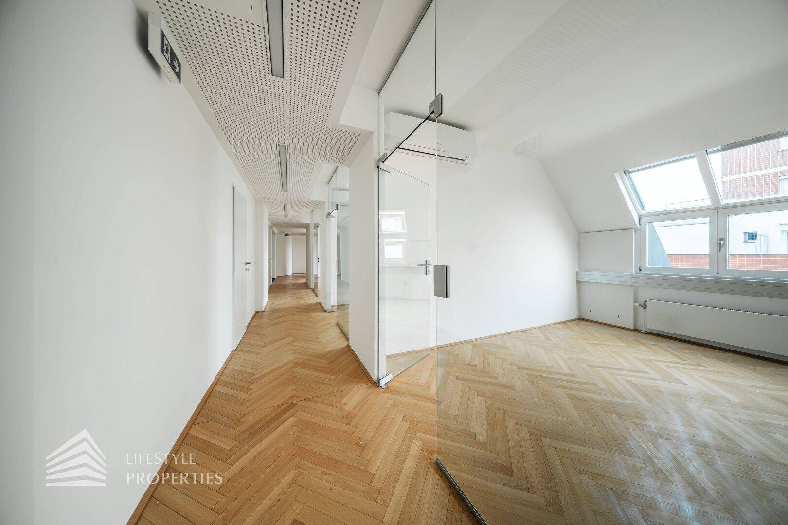 Langer Bürogang mit Glaswand, Fischgrätparkett und perforierter Decke, führt zu weiteren hellen Räumen.
