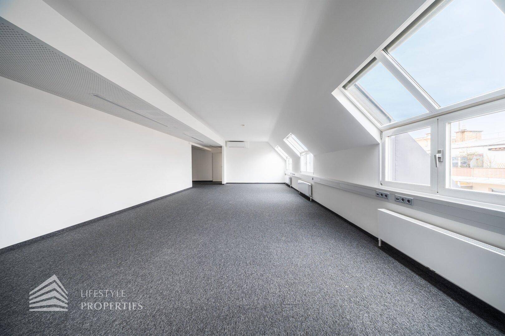Weitläufiger Bürobereich mit schrägen Wänden, Teppichboden und viel Tageslicht.