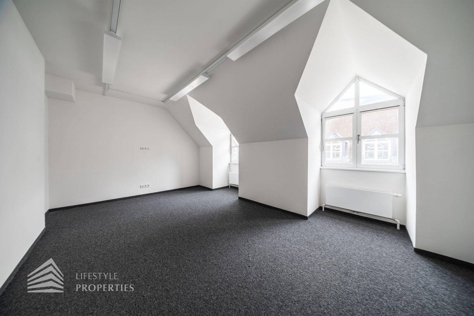 Heller Büroraum mit Teppichboden, schrägen Wänden und einem markanten Dachfenster.