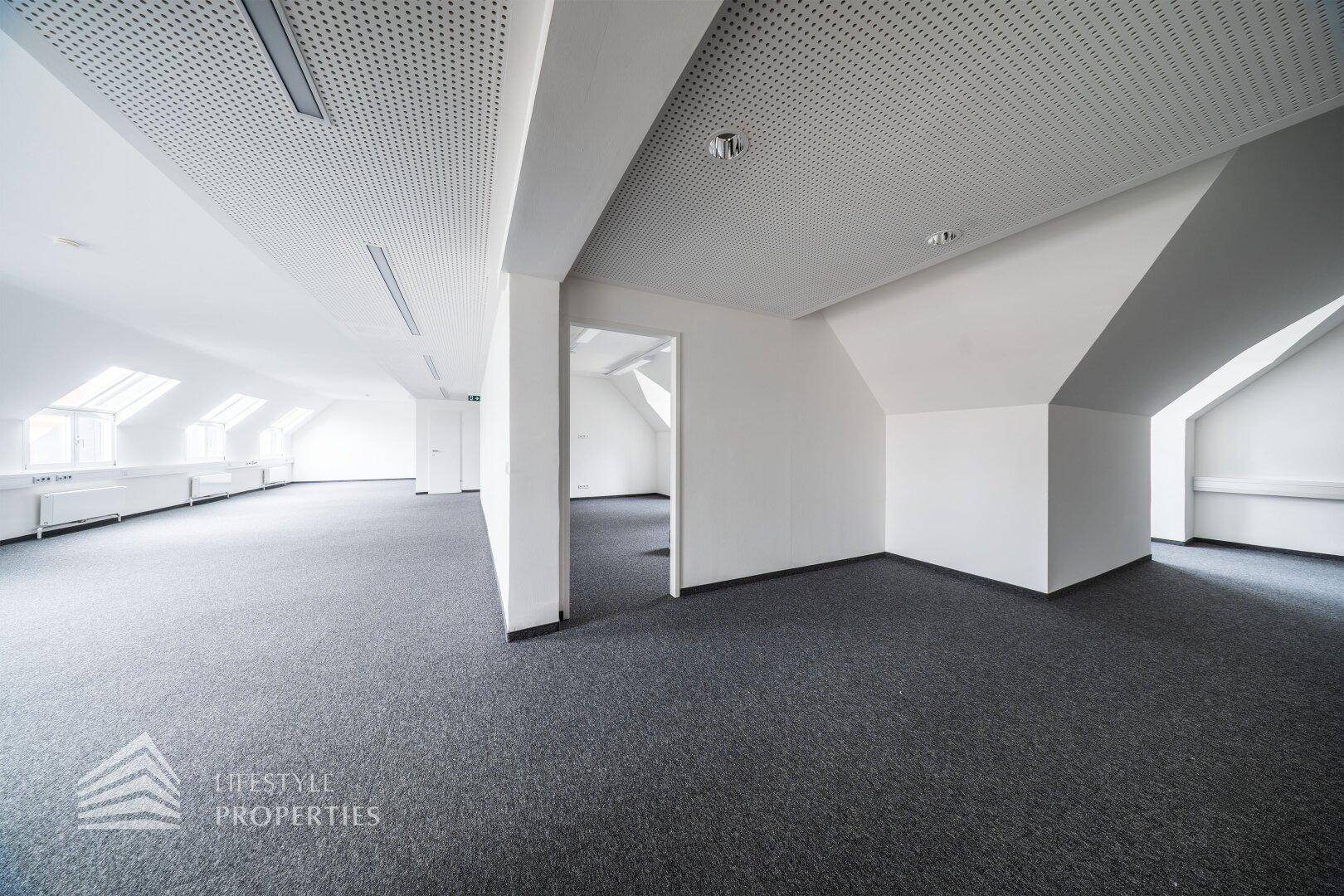Weitläufiger Bürobereich mit Teppichboden, schrägen Wänden und offener Raumgestaltung.