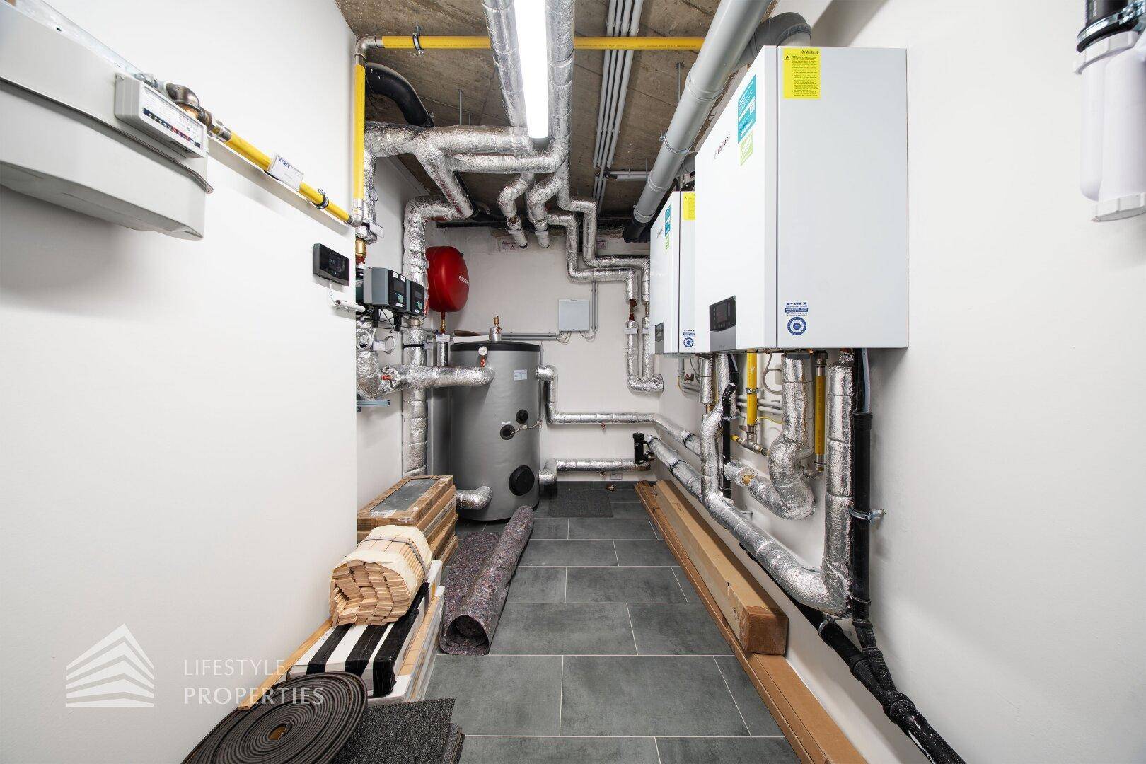 Technikraum mit Heizungsanlagen, Warmwasserspeicher und zahlreichen Rohren an Wänden und Decke.