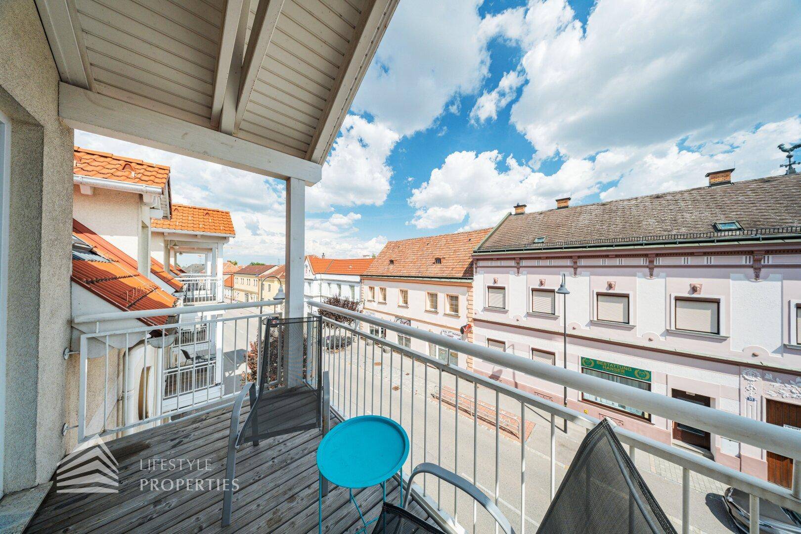 Blick von einem möblierten Balkon auf eine belebte Straße mit traditionellen Gebäuden unter blauem Himmel.