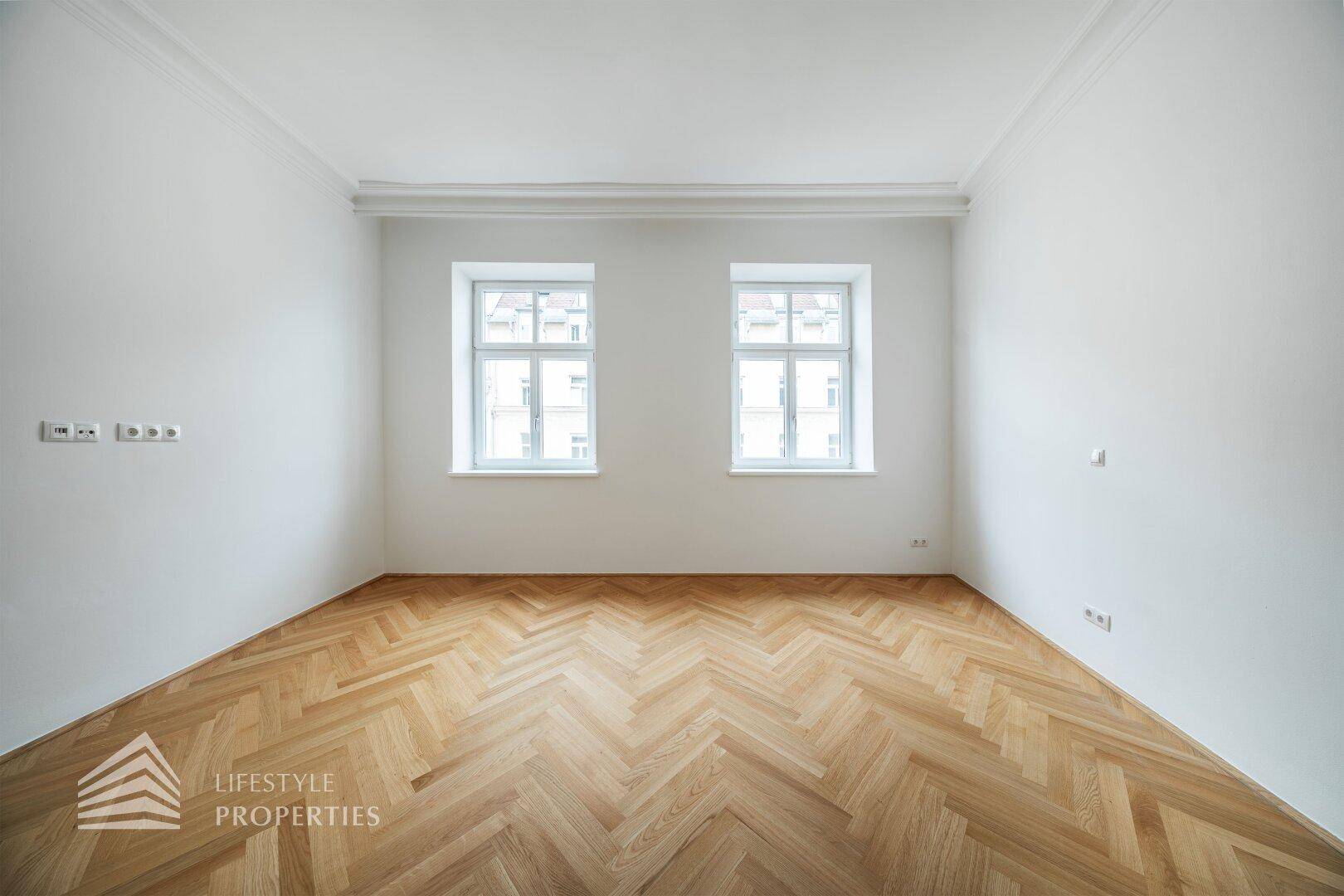 Heller, leerer Raum mit zwei Fenstern, hohen Decken und elegantem Fischgrätparkettboden.
