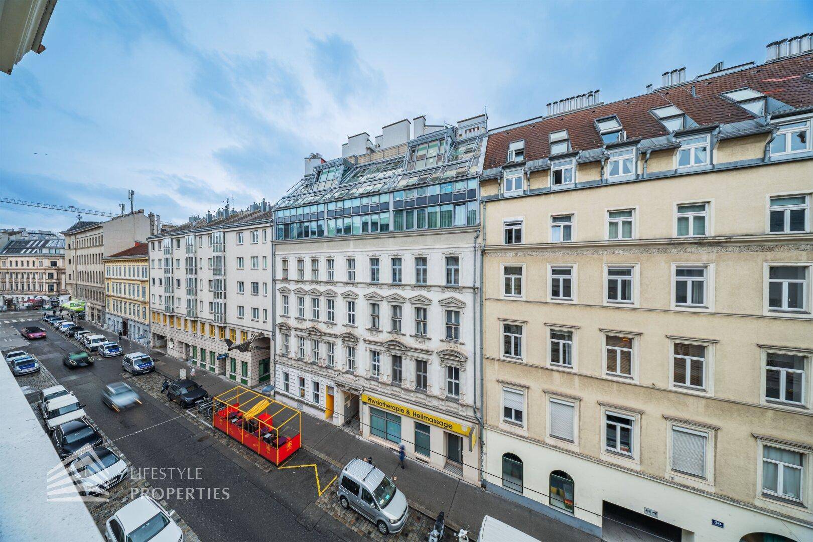 Blick von oben auf eine belebte Stadtstraße mit historischen Gebäuden und modernen Dächern.