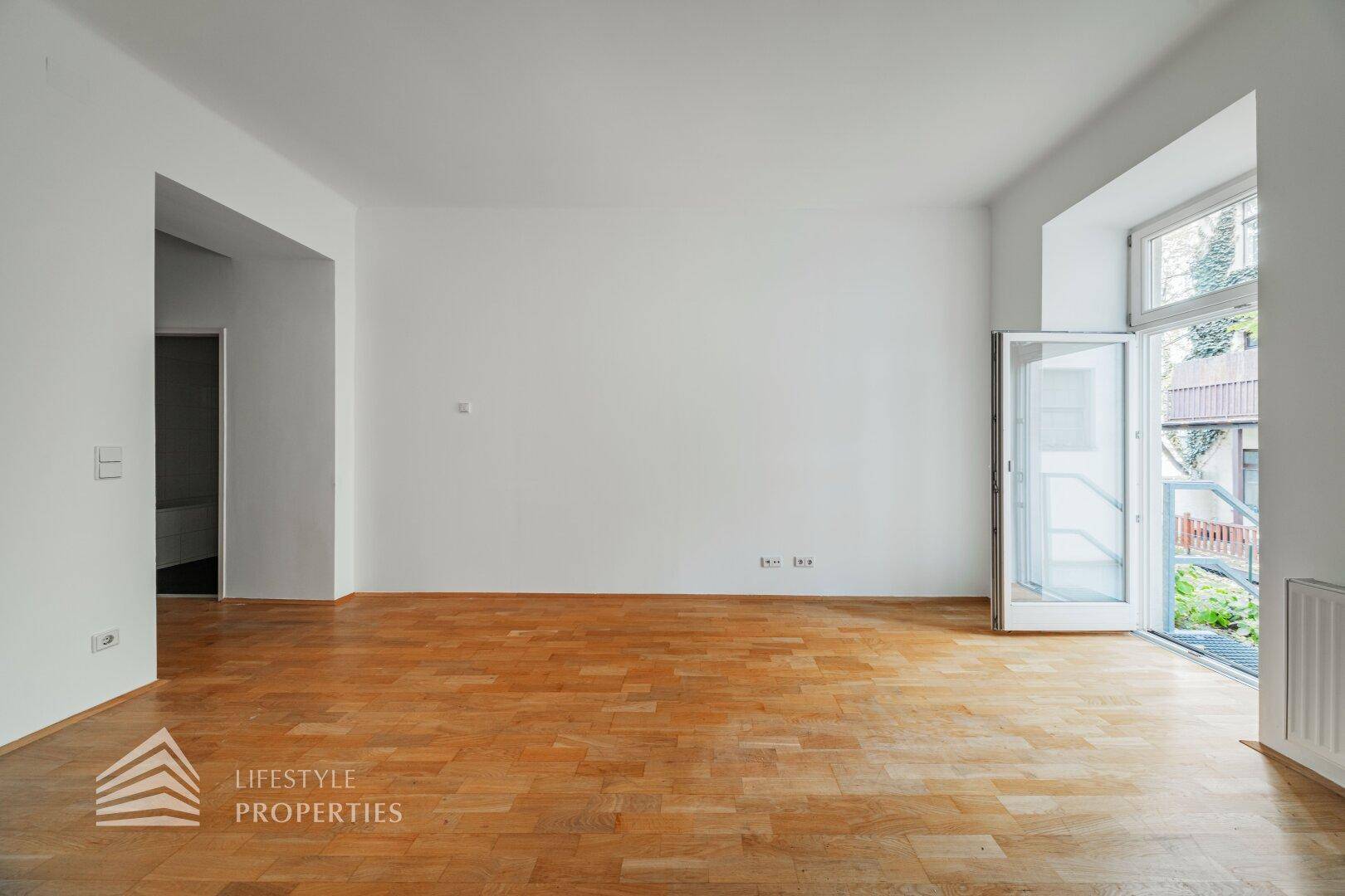 Leerer Wohnraum mit Parkettboden und Zugang zu einem kleinen Außenbereich, bereit zur individuellen Gestaltung.