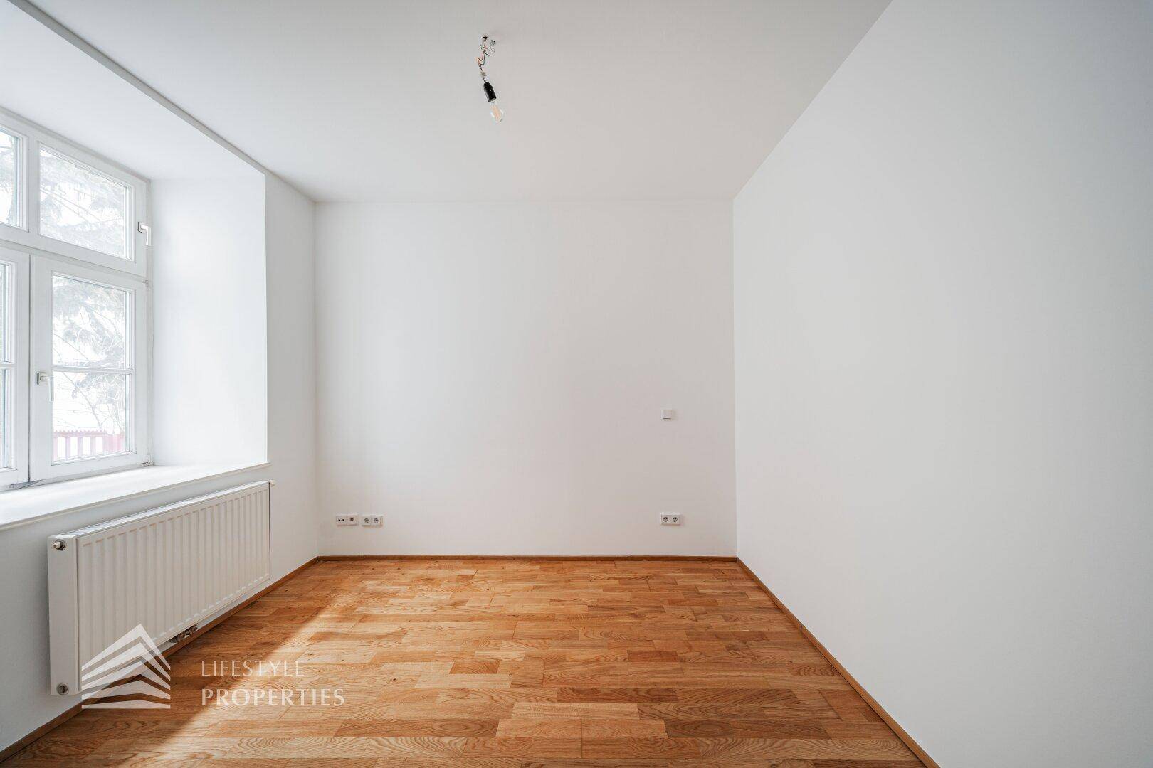 Leeres Zimmer mit Parkettboden und großem Fenster, ideal als Schlafzimmer oder Arbeitsbereich.