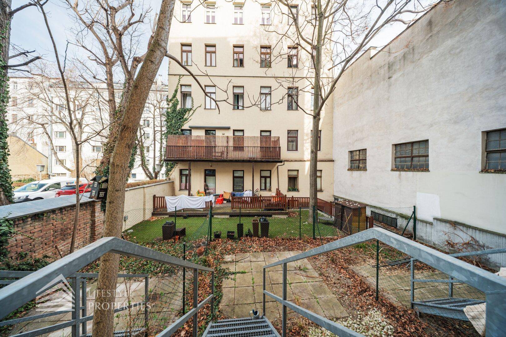 Blick auf einen kleinen Gartenbereich mit Rasen und Balkonen im Hintergrund, umgeben von Bäumen.