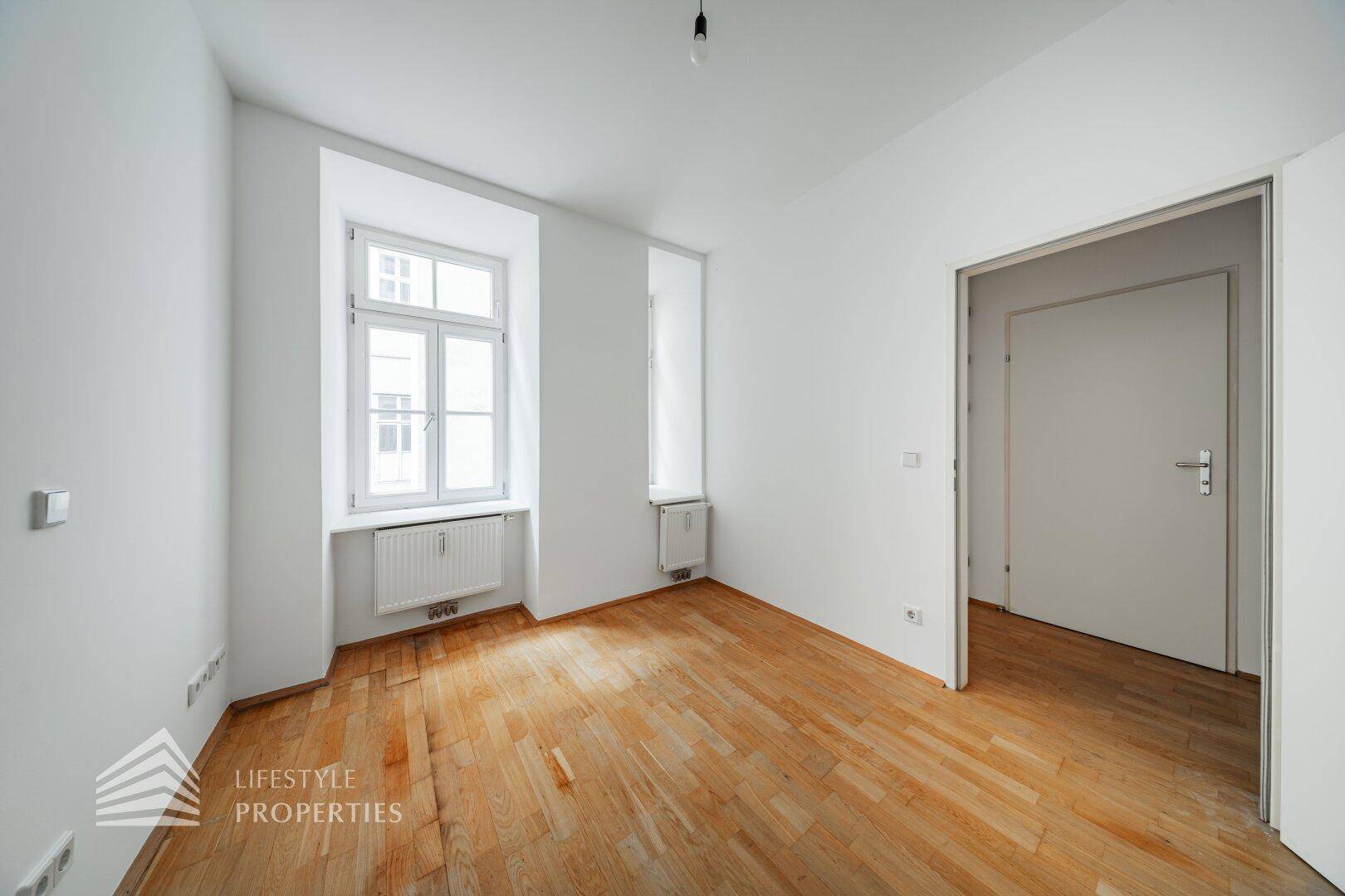 Leeres Zimmer mit Parkettboden und großem Fenster, ideal als Schlaf- oder Arbeitszimmer.