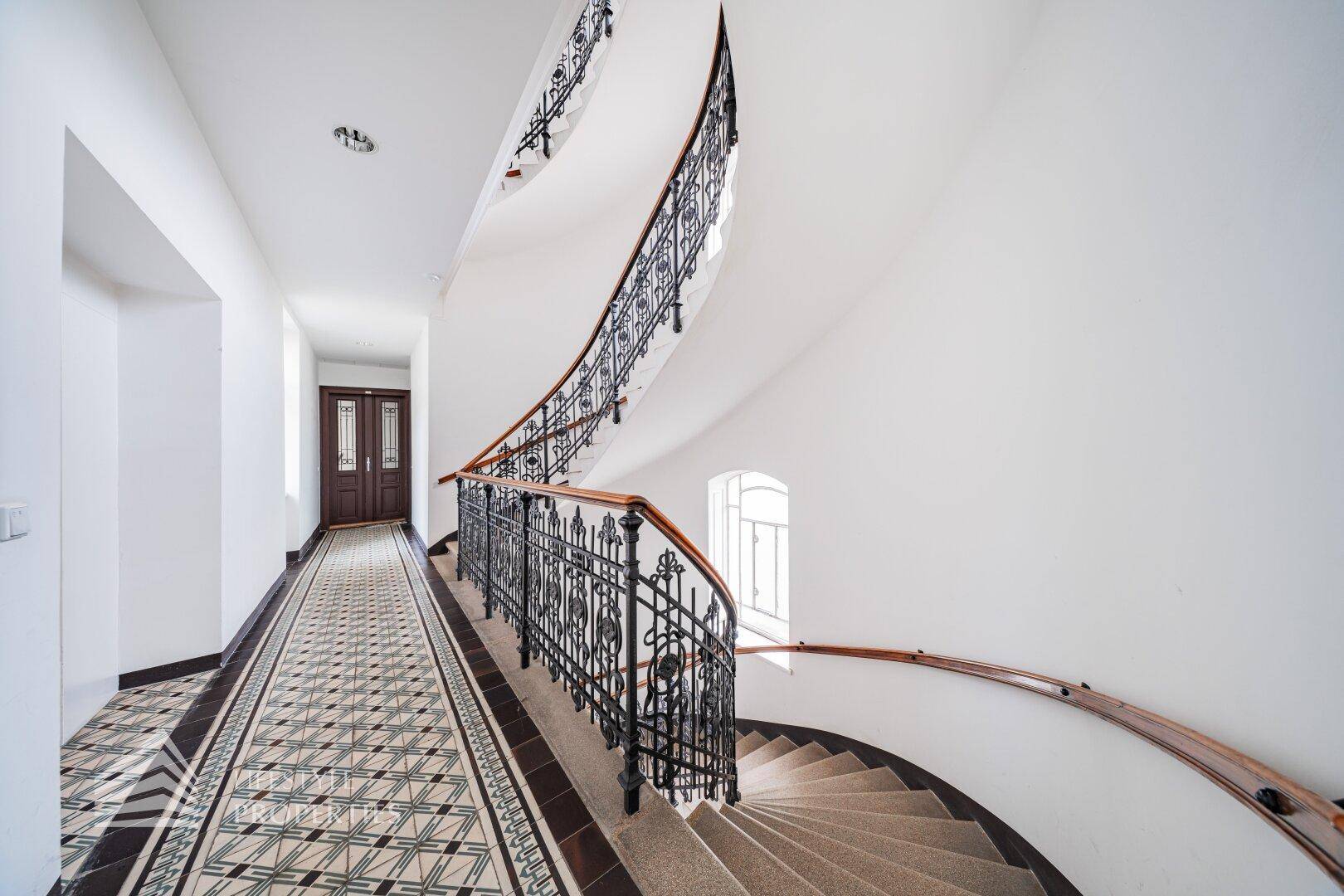 Elegantes Treppenhaus mit geschwungenem Geländer, gemusterten Fliesen und viel Tageslicht.