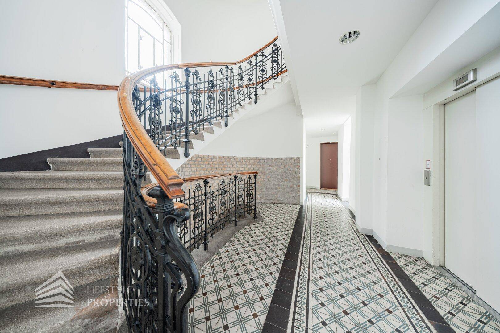 Elegantes Treppenhaus mit schmiedeeisernem Geländer, Holzakzenten und gemustertem Fliesenboden.