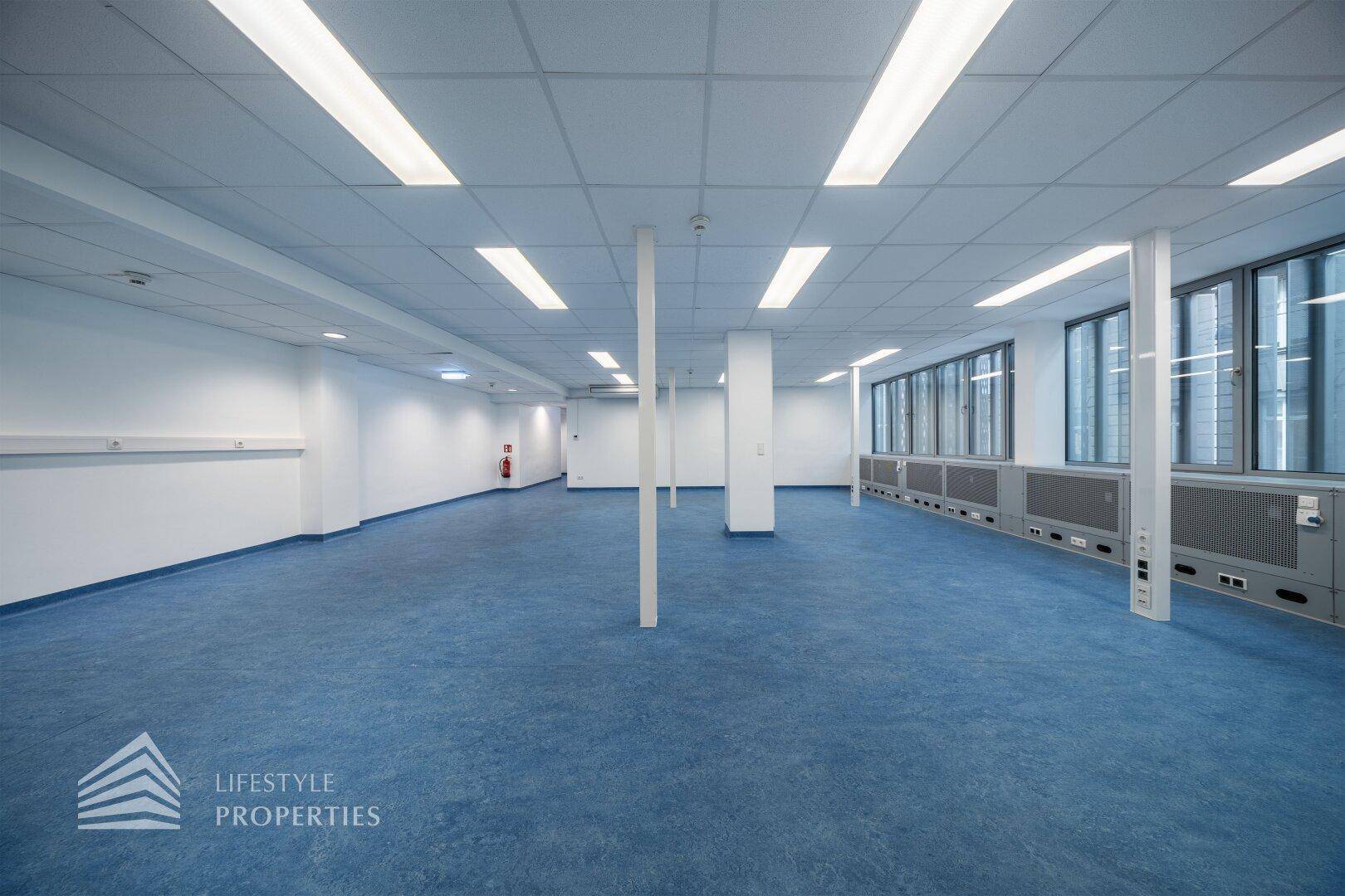 Weitläufiger Büroraum mit blauen Bodenbelag, weißen Säulen und einer Fensterfront, die viel Licht spendet.