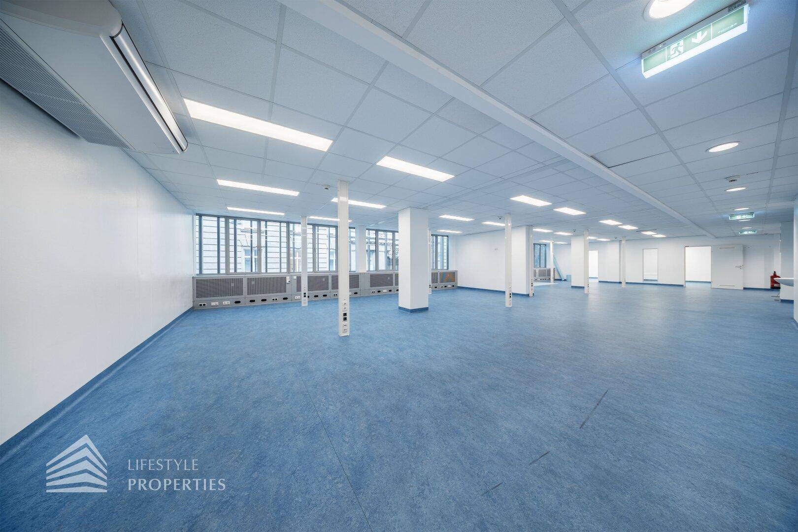 Großer, offener Bürobereich mit blauen Bodenbelag, vielen Fenstern und einer Klimaanlage an der Decke.