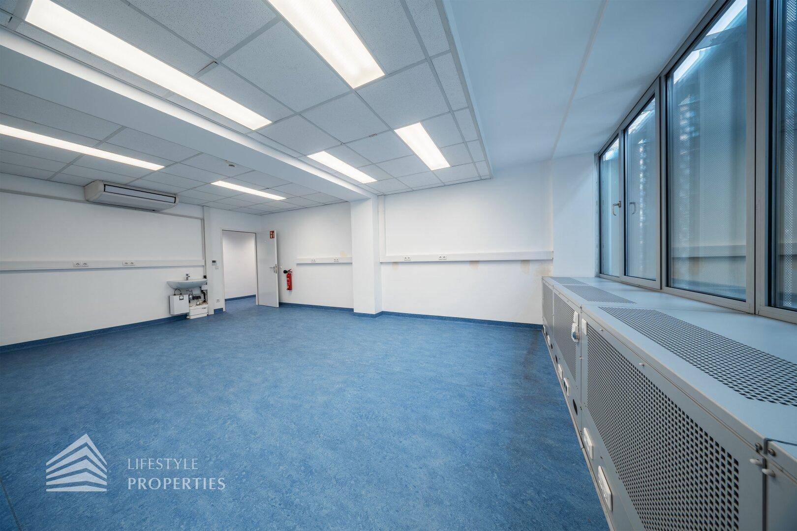 Leerer Büroraum mit blauen Bodenbelag, großen Fenstern und einem Waschbecken in der Ecke.