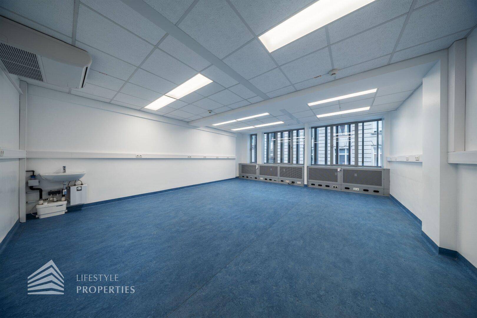 Geräumiger Büroraum mit blauen Bodenbelag, großen Fenstern und einem Waschbecken an der Wand.