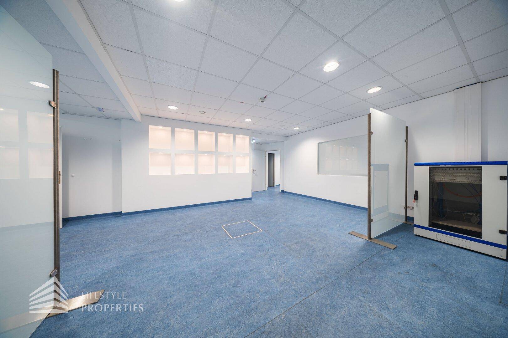 Offener Bürobereich mit blauen Bodenbelag, Glaswänden und einer beleuchteten Nische in der Wand.