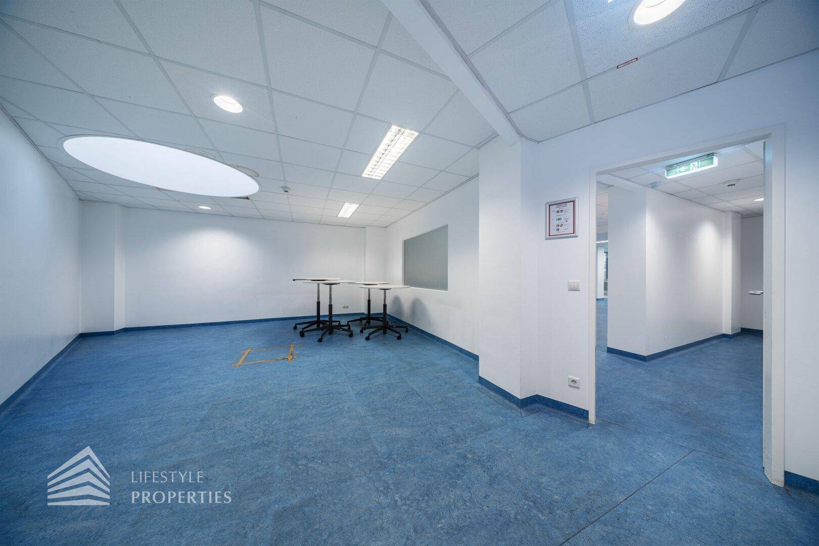 Büroraum mit blauen Bodenbelag, runder Deckenöffnung und mobilen Stehtischen in der Mitte.