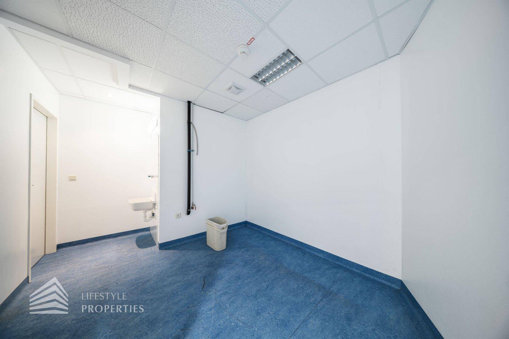 Kleiner Büroraum mit Waschbecken, blauen Bodenbelag und weiß gestrichenen Wänden.