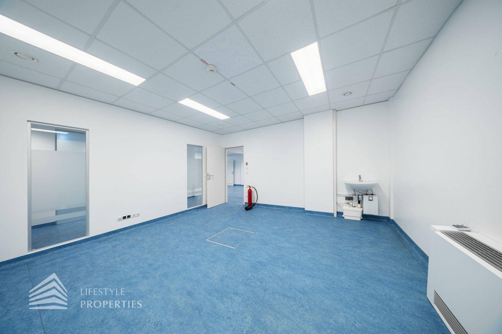 Büroraum mit blauen Bodenbelag, Waschbecken und einem Feuerlöscher an der Wand.
