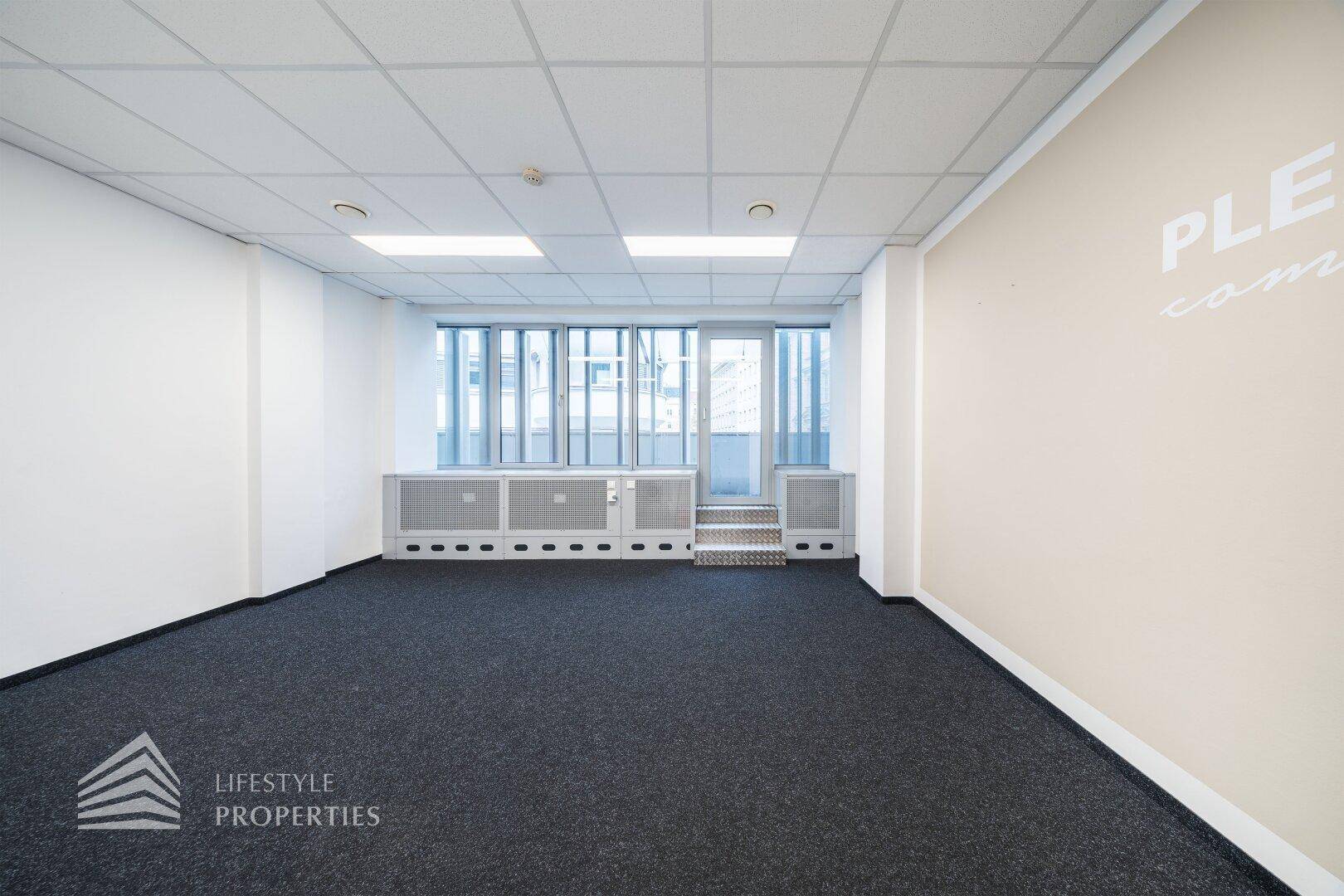 Bürofläche mit Teppichboden, einer hellbeigen Akzentwand und Fenstern, die den Raum mit Tageslicht füllen.