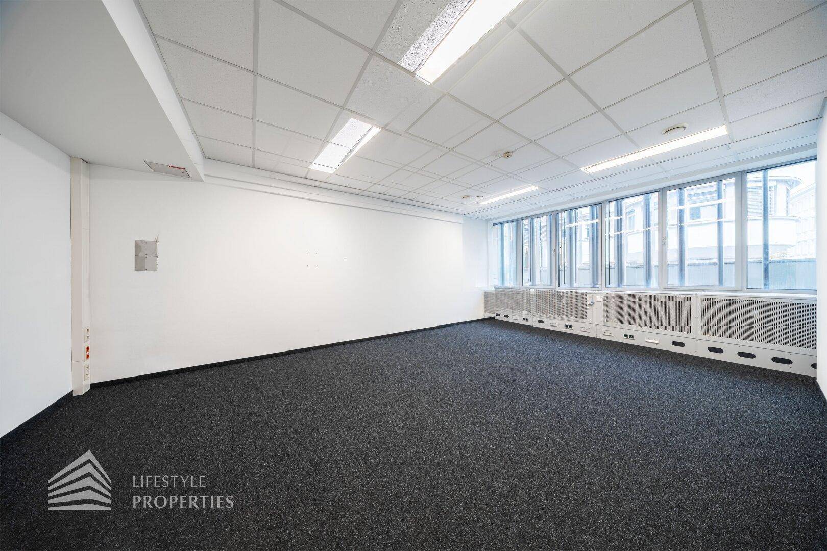 Leerer Büroraum mit Teppichboden, weißen Wänden und einer Fensterfront, die viel natürliches Licht spendet.