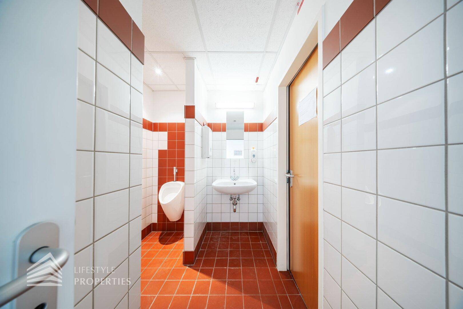 Sanitärbereich mit weißen und roten Fliesen, einem Urinal und einem Waschbecken.