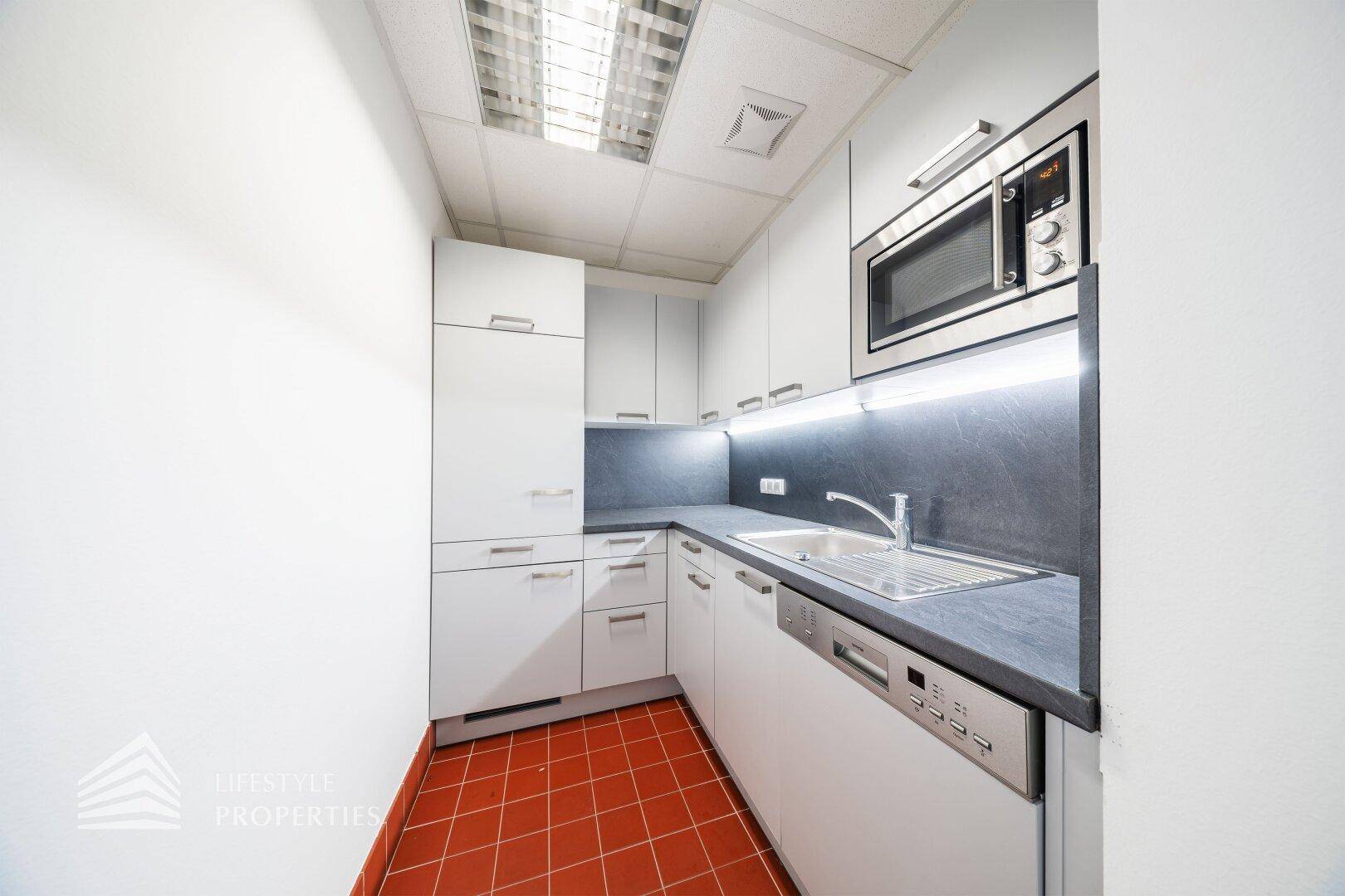 Kleine, moderne Teeküche mit weißen Schränken, dunkler Arbeitsplatte und roten Fliesenboden.