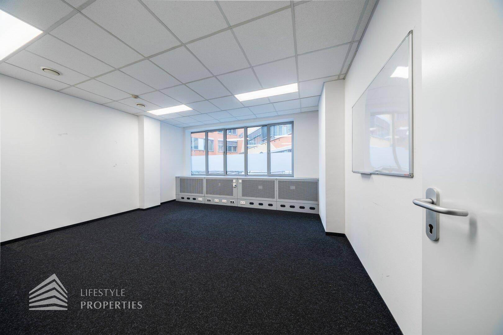 Leeres Büro mit Teppichboden, weißen Wänden und einem Fenster, das Tageslicht in den Raum bringt.