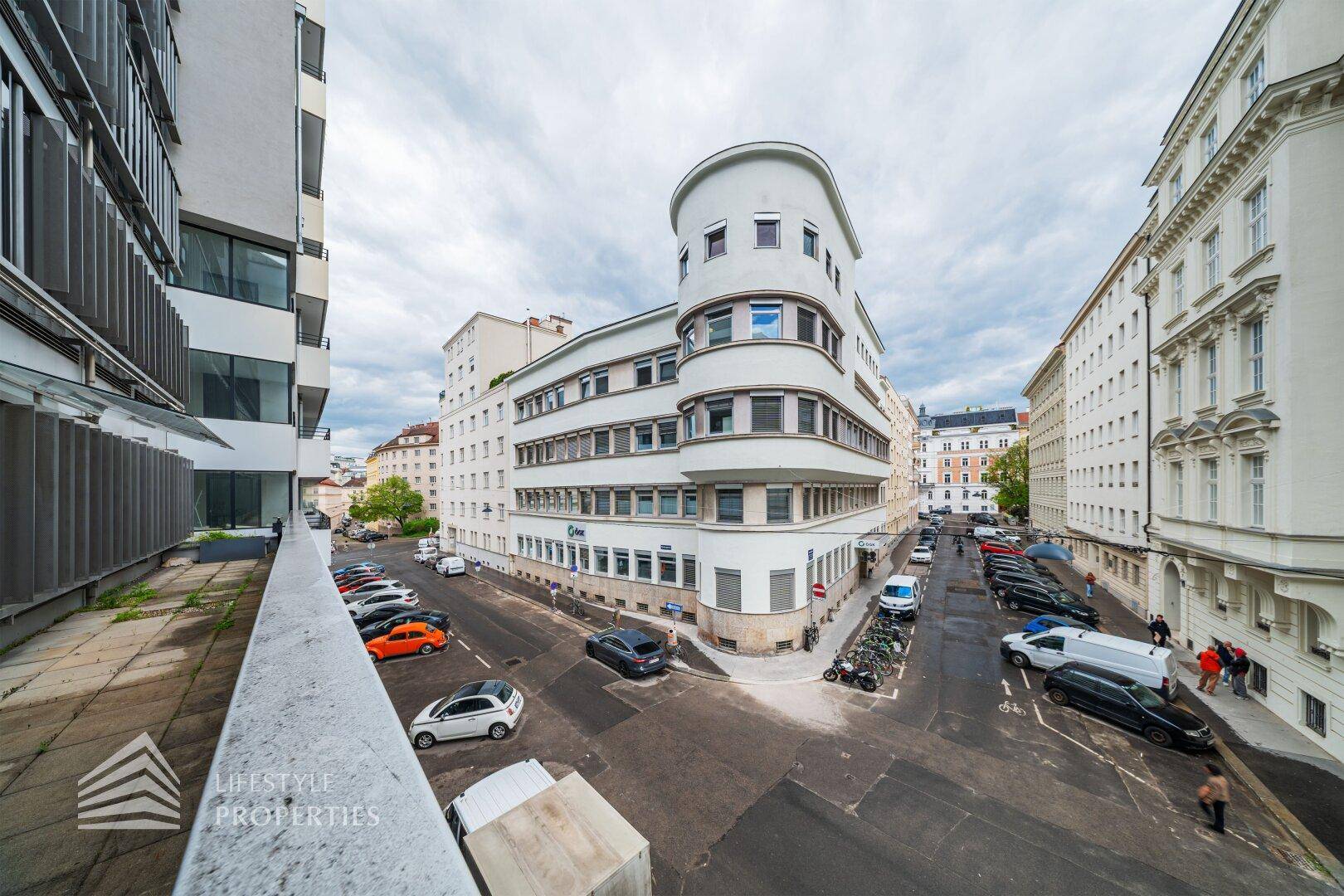 Panoramablick von der Terrasse auf eine städtische Kreuzung mit verschiedenen Gebäudestilen und parkenden Fahrzeugen.