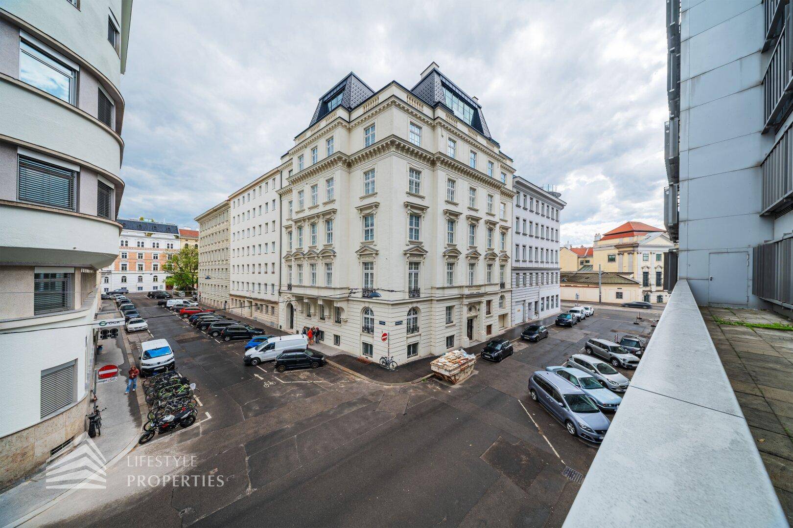 Blick von der Terrasse auf eine belebte Stadtstraße mit parkenden Autos und historischen Gebäuden.