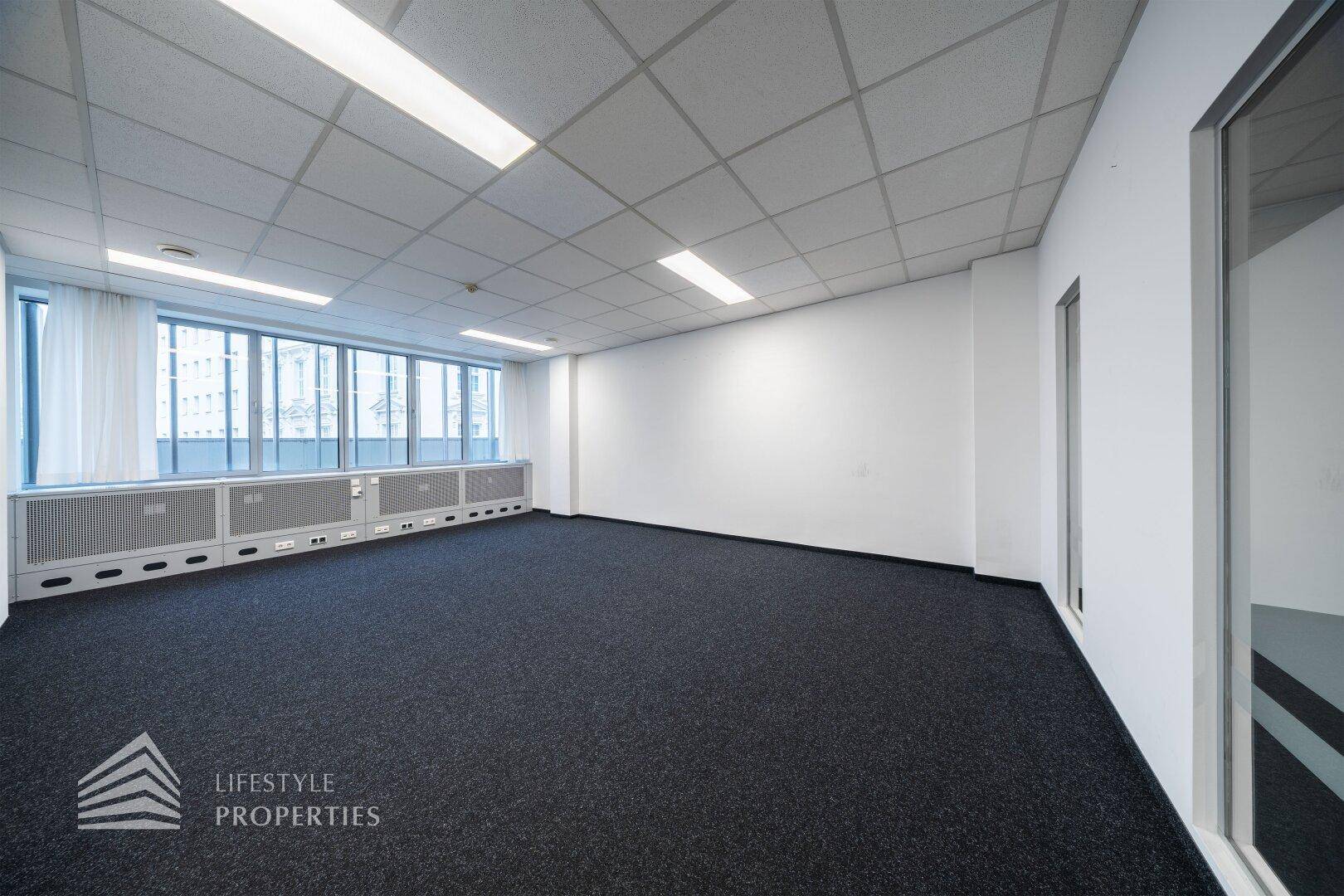 Leeres Büro mit Teppichboden, weißen Wänden und großen Fenstern, die für eine helle Atmosphäre sorgen.