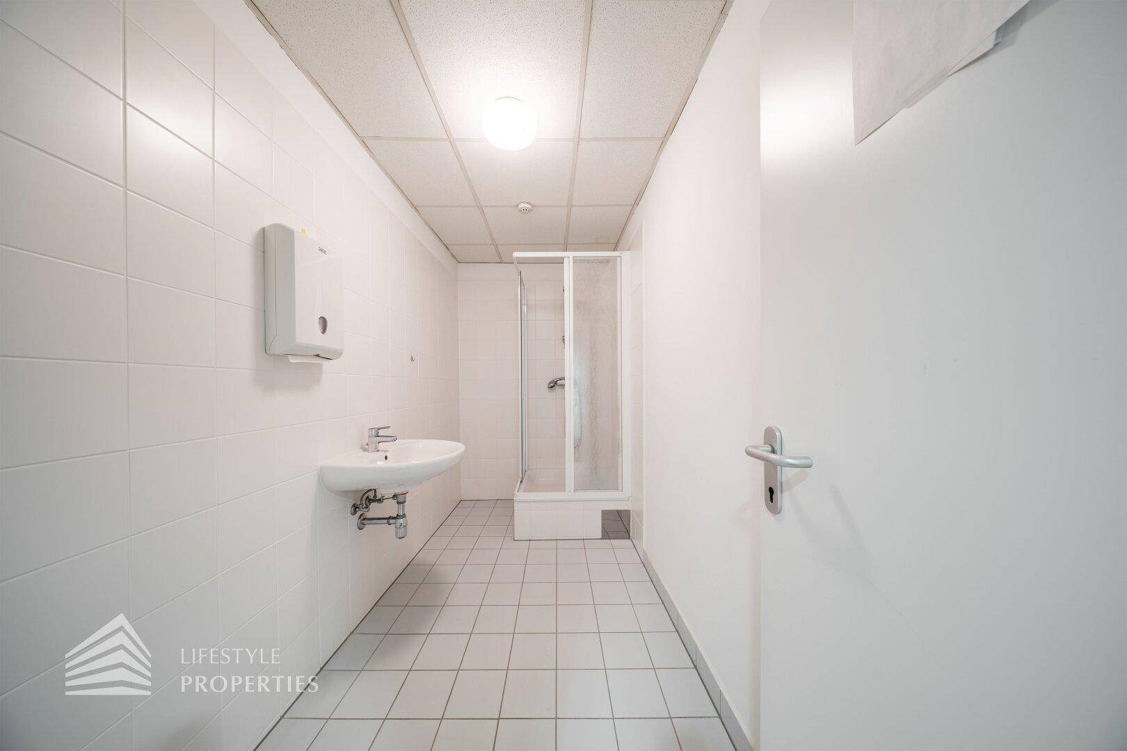 Heller Sanitärraum mit Dusche und Waschbecken, ausgestattet mit weißen Fliesen und Spender.