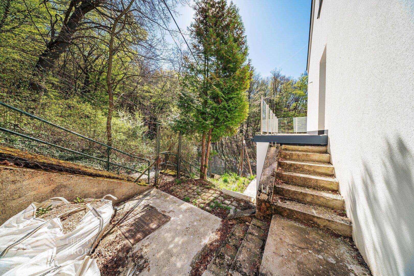 Außenbereich mit Treppen und Baustellenresten, umgeben von Bäumen und viel Grün.