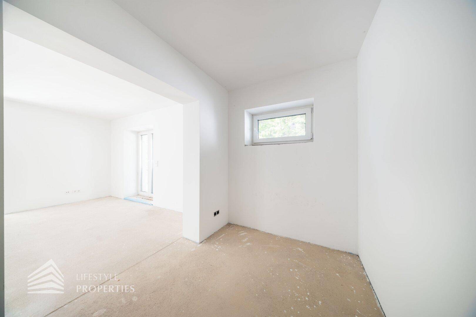 Heller Raum im Rohbau mit Fenster und offenem Durchgang zu einem weiteren Bereich.