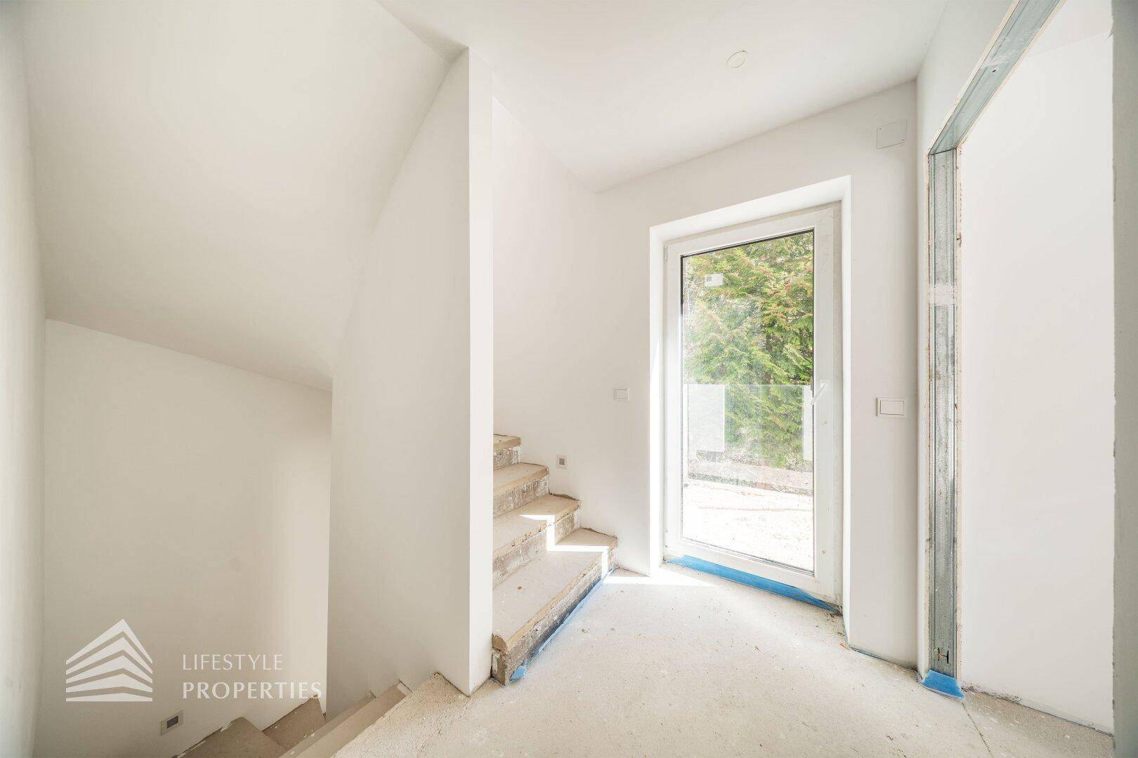 Treppenaufgang im Rohbau mit hellem Fenster und Zugang zum Außenbereich, lichtdurchflutet.