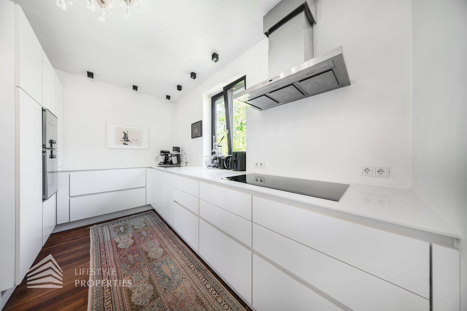Moderne weiße Küche mit Einbaugeräten, Induktionskochfeld und Fenster mit Blick ins Grüne.
