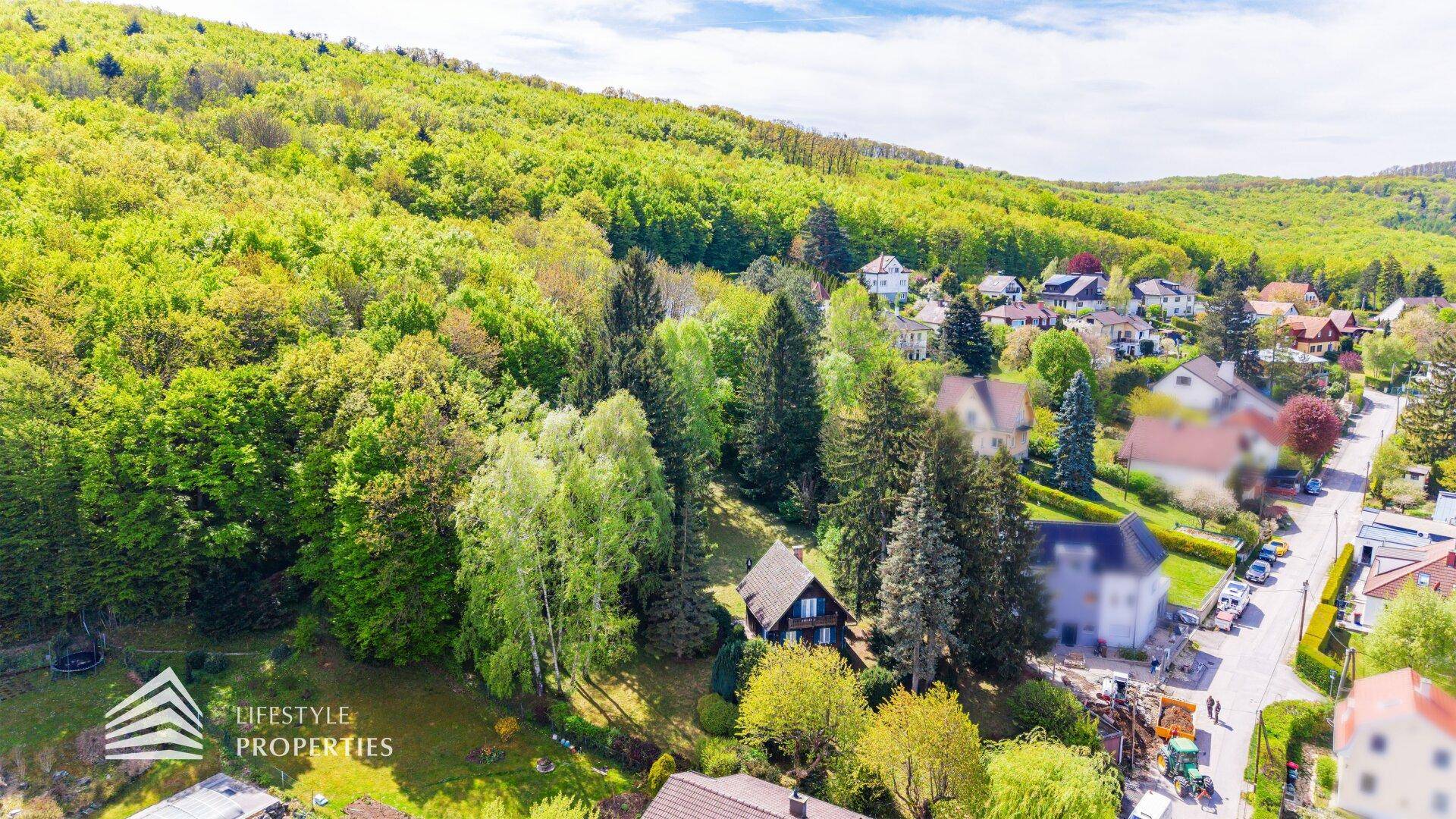 Weitläufige Luftaufnahme des Holzhauses, eingebettet in eine grüne Waldlandschaft mit angrenzender Siedlung.