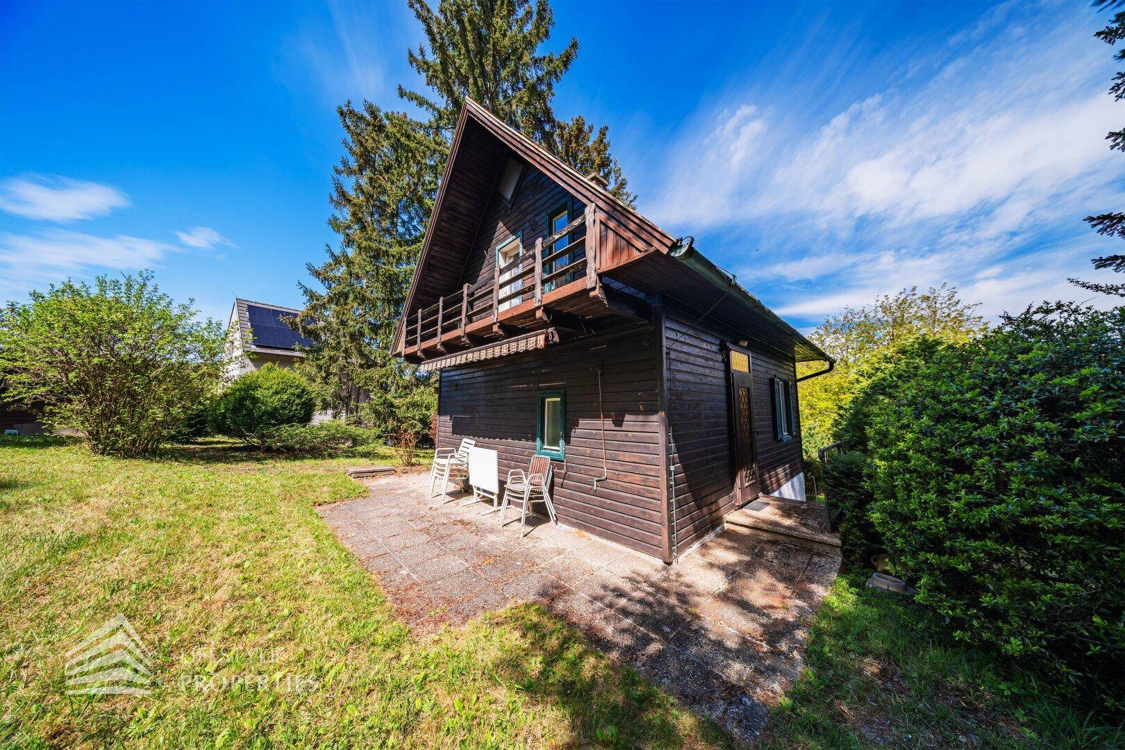 Charmantes Holzhaus mit kleiner gepflasterter Terrasse und Gartenmöbeln, umgeben von viel Grün.