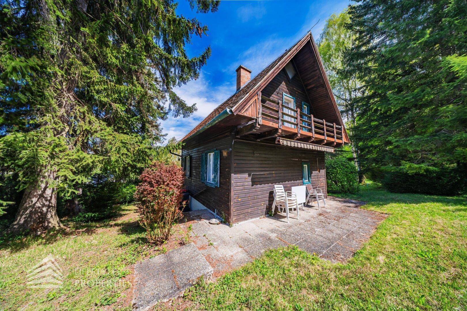 Rustikales Holzhaus mit kleiner Terrasse und Gartenmöbeln, umgeben von hohen Bäumen und Natur.