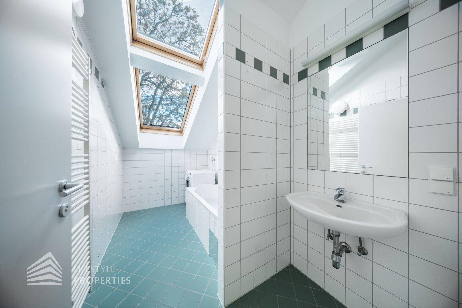 Helles Badezimmer mit weißen Wandfliesen, blauen Bodenfliesen, Waschbecken und Dachfenster.