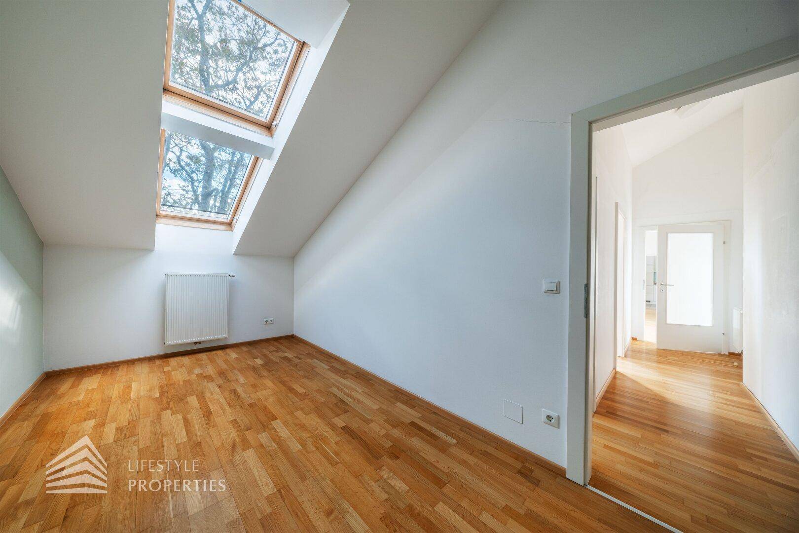 Helles Schlafzimmer mit Parkettboden und Dachfenstern, das zu einem Flur führt.