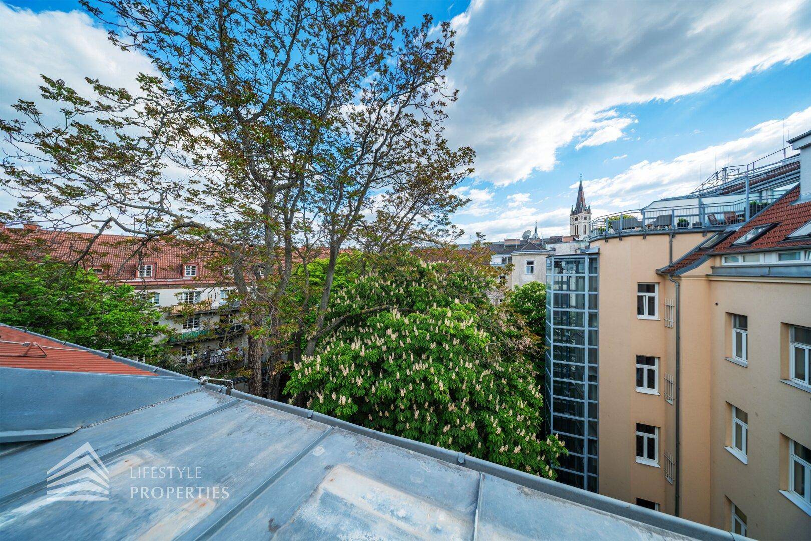 Panoramablick über die Dächer der Stadt mit viel Grün und einem Kirchturm im Hintergrund.