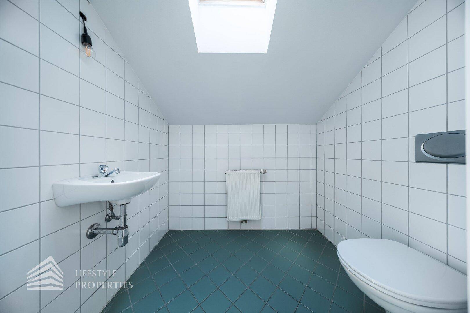 Separates WC mit Waschbecken, Wand-WC und weißen Fliesen, beleuchtet durch ein Dachfenster.