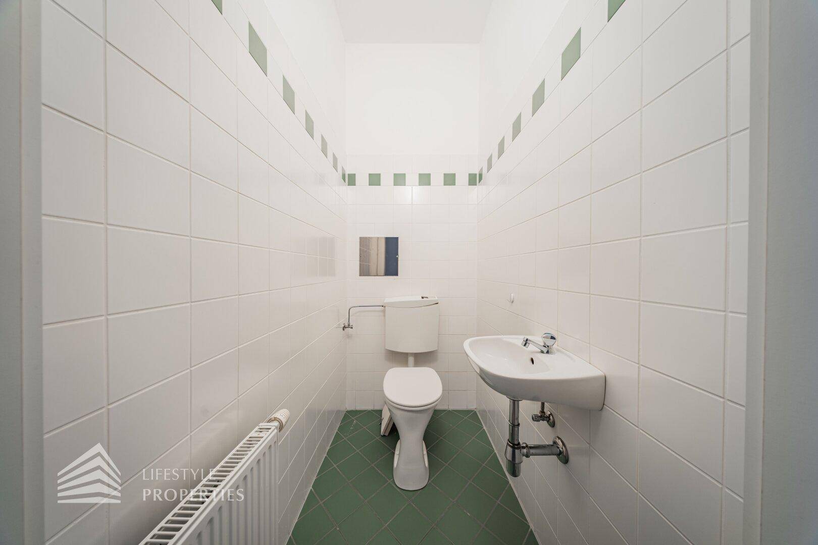 Separates WC mit Toilette, Waschbecken und weißen Fliesen mit grünen Akzentstreifen.