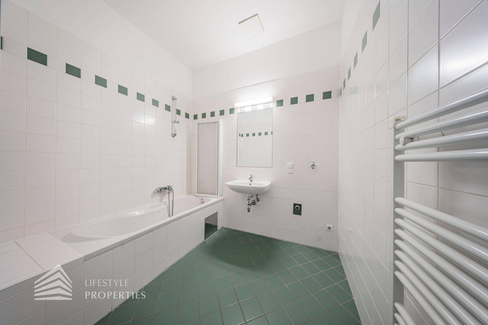 Geräumiges Badezimmer mit Badewanne, Waschbecken und weißen Fliesen mit grünen Akzenten.